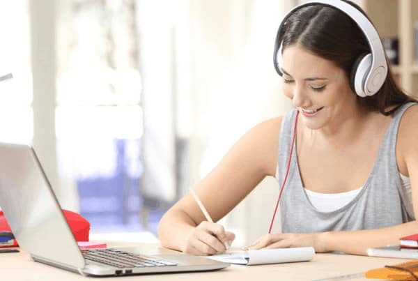 student with headphones writing notes with open laptop