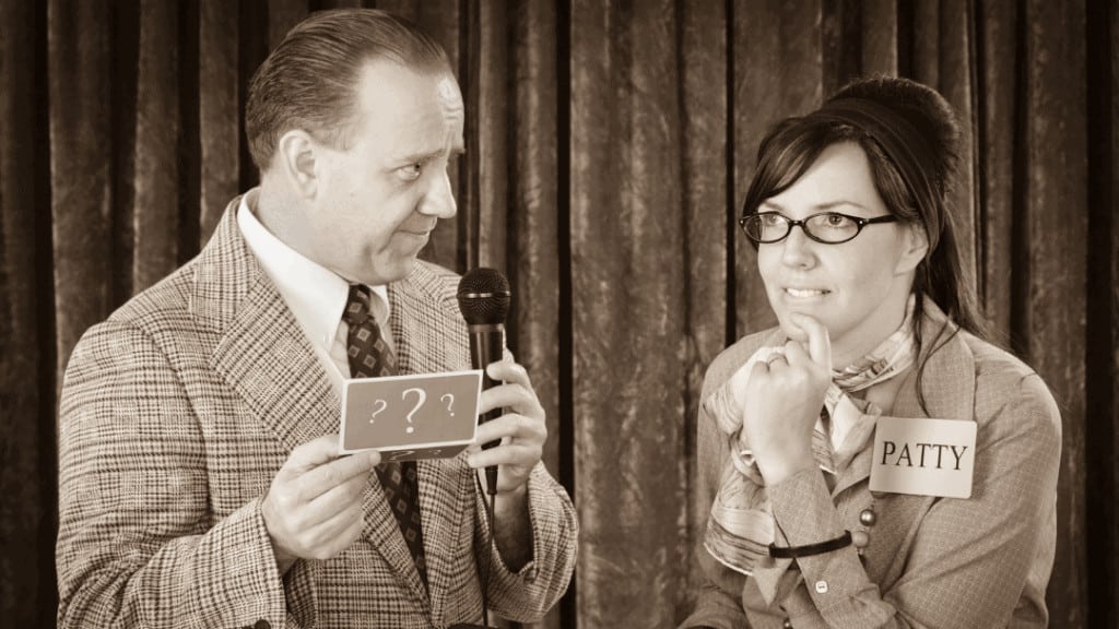 a man on the left in a suit jacket and tie holding a microphone and card with question marks, a woman with glasses, scarf and name tag "Patty" thinking with finger on her chin