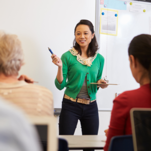 female educator in front of whiteboard talks to adult students in chairs